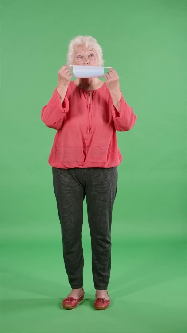 Ritratto anziana donna guardando la macchina fotografica e mette su maschera medica. Nonna dai capelli grigi in camicetta rossa su schermo verde a studio. Al rallentatore. Video verticale. — Video Stock