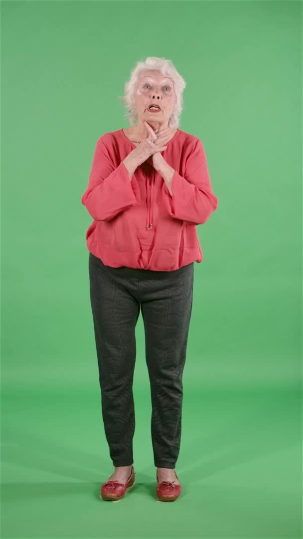 Портрет пожилой женщины, смотрящей в камеру, держащей ладонь вместе и просящей чего-то. Серая бабушка в красной блузке на зеленом экране в студии. Медленное движение. Вертикальное видео. — стоковое видео