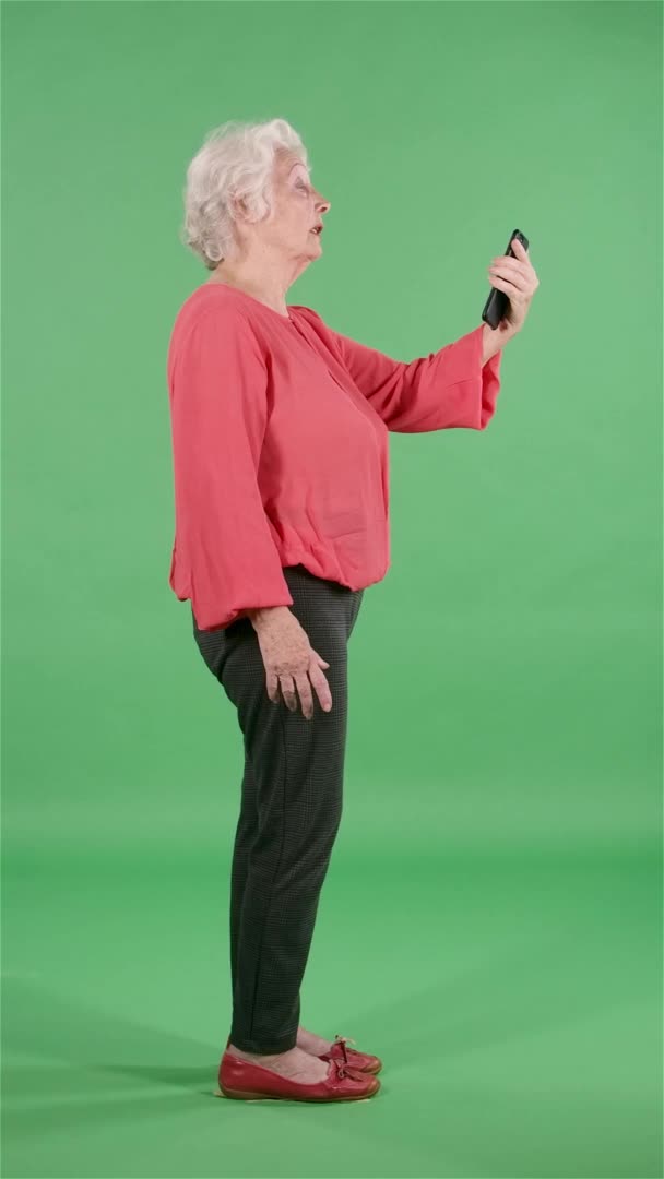 Портрет пожилой женщины, разговаривающей по видеосвязи со своего смартфона. Серая бабушка в красной блузке на зеленом экране в студии. Вид сбоку. Медленное движение. Вертикальное видео. — стоковое видео