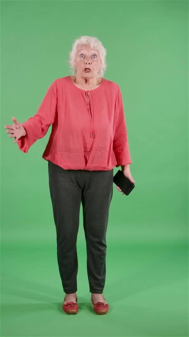 Portrait elderly woman looking at the camera is emotionally talking to someone with indignation. Gray haired grandmother in red blouse on green screen at studio. Slow motion. Vertical video. — Stock Video