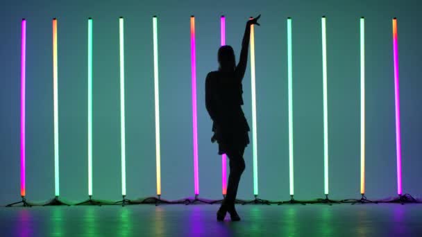 En ung kvinna visar element av rumba dans i studion mot bakgrund av flerfärgade neonrör. Passionerad latinamerikansk dans i slow motion. — Stockvideo