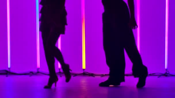 Penari profesional menari rumba di studio dengan latar belakang tabung neon terang. Tutup penari kaki berlatih langkah-langkah tari. Siluet. Gerakan lambat. — Stok Video