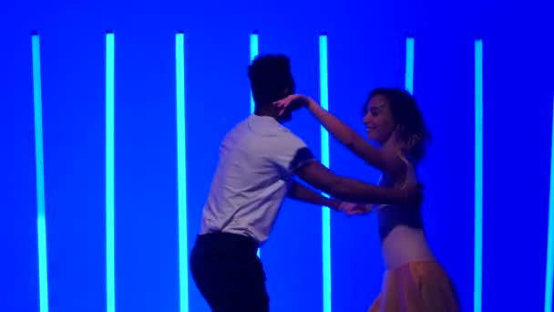 Flexibele en sensuele dansers oefenen een reeks snelle draaibeurten tegen een achtergrond van felblauwe neonlichten in een donkere studio. Glimlachend meisje wervelt hand in hand met haar partner. Langzame beweging. Sluiten.. — Stockvideo