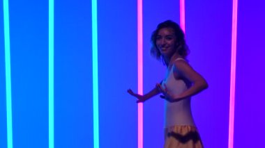 İnce dansçı, salsa dansının hareketlerini çalışırken kalçalarını hareket ettirir. Bir kadın stüdyoda yavaş çekimde mavi ve pembe neon ışıkların arka planına karşı dans ediyor. Kapat..