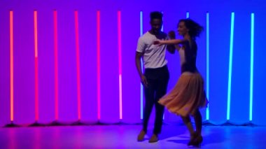 Mutlu siyah adam ve beyaz kadın, arka planda çok renkli neon ışıkların altında dönüp duruyorlar. Tutkulu Latin Amerika salsası yavaş çekimde dans ediyor.