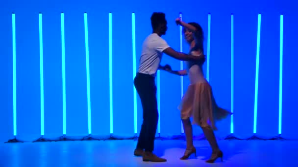 Los bailarines flexibles y sensuales practican una serie de giros rápidos contra un telón de fondo de luces de neón azul brillante en un estudio oscuro. Chica sonriente está girando cogida de la mano con su pareja. Movimiento lento. — Vídeos de Stock