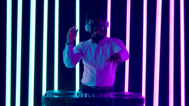Портрет молодого афроамериканського чоловіка з великими білими навушниками, який танцює і полюбляє змішувати музику на ігровому столі ді-джея. Повільний рух. — стокове відео