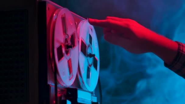 Man hand rotates reels on old analogue reel to reel audio tape recorder. Vintage music player on a blue smoky studio background with red neon lights. Close up. Slow motion. — Stock Video