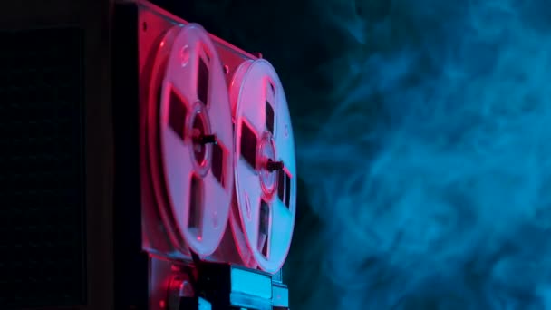 Man hand stops playing old analogue reel to reel audio tape recorder. Vintage music player on a blue smoky studio background with red neon lights. Close up. Slow motion. — Stock Video