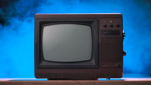 Vintage televisie tegen een rokerige studio achtergrond met blauwe neon lichten en stofdeeltjes. Oude TV staat op houten tafel. Retro atmosfeer. Langzame beweging. — Stockvideo