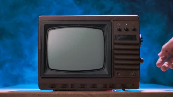 El hombre trata de encender el viejo televisor, golpea contra él, dispersando partículas de polvo. Televisión vintage sobre un fondo de estudio ahumado con luces de neón azul. Las manos masculinas se cierran. Movimiento lento. — Vídeo de stock