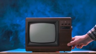 Adam eski televizyonu açmaya çalışıyor, kapıyı çalıyor, toz parçacıkları saçıyor. Smoky stüdyo arka planına karşı mavi neon ışıkları olan klasik televizyon. Erkek elleri yakın. Yavaş çekim.
