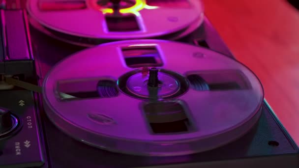 鮮やかなクラブネオンライトに照らされたヴィンテージテープレコーダー上のレトロ磁気テープスピンの透明なリール。古い音楽はレトロなパーティーで演奏します。70 、 80年のためのノスタルジア。閉めろ。スローモーション — ストック動画