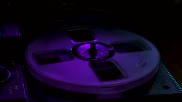 男性の手は紫とピンクのネオンライトで照らされたリールテープレコーダーにヴィンテージプロアナログをオンにします。透明な回転コイルは古い音楽を再現します。レトロなテープだ。閉めろ。スローモーション. — ストック動画