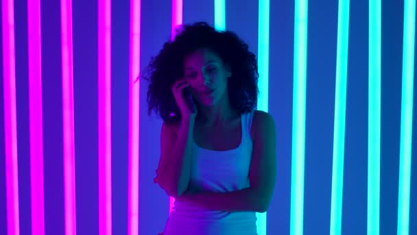 Portrait of a young African American woman talking on the phone. Black woman with curly hair poses in light of multicolored neon tubes. Close up. Slow motion. — Stock Video