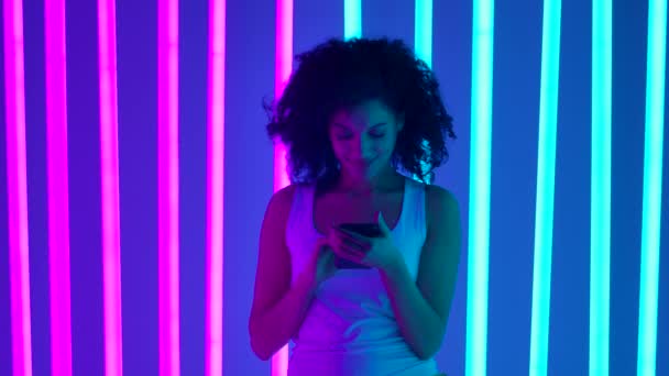 Portrait d'une jeune belle femme qui écrit des SMS sur son téléphone avec un sourire énigmatique. Une belle fille pose dans le studio entouré de tubes néons rose vif et bleu. Mouvement lent. — Video