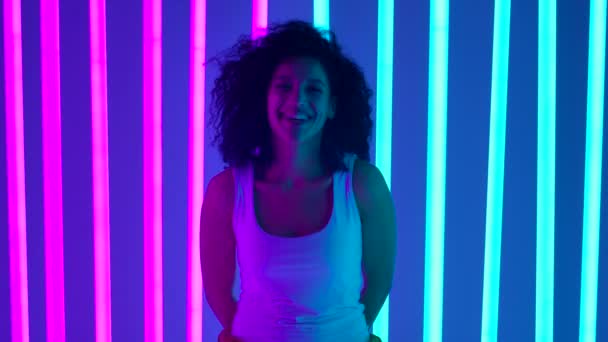 Egy fiatal afro-amerikai nő portréja, aki a kamerába néz és mosolyog. Egy bájos lány pózol a stúdióban színes neoncsöveken. Lassú mozgás. Közelről.. — Stock videók