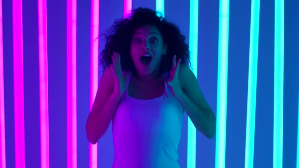 A African American woman holds her fists in anticipation, is happily surprised and says wow, claps her hands. Girl against a background of blue and pink neon lights. Slow motion. Close up. — Stock Video