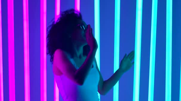 Jovem afro-americana em óculos de sol elegantes dançando no estúdio contra um fundo de luzes de néon azul e rosa. Clube nocturno. Movimento lento. Fechar. — Vídeo de Stock