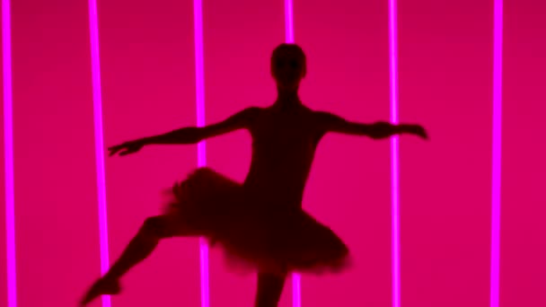 Ung ballerina i tåskor och vit baletttutu gör en piruett. Repetition av den vita svandelen i en mörk ateljé mot bakgrund av starkt neonljus. Stäng siluetten. Långsamma rörelser. — Stockvideo