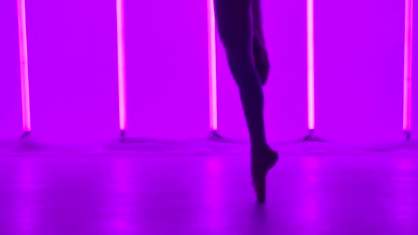 Ballerina framför piruett medan dansande inslag av klassisk balett i en mörk ateljé. Ben kvinnliga dansare i tåskor på tå mot en bakgrund av ljusa neon ljus närbild. Närbild. — Stockvideo