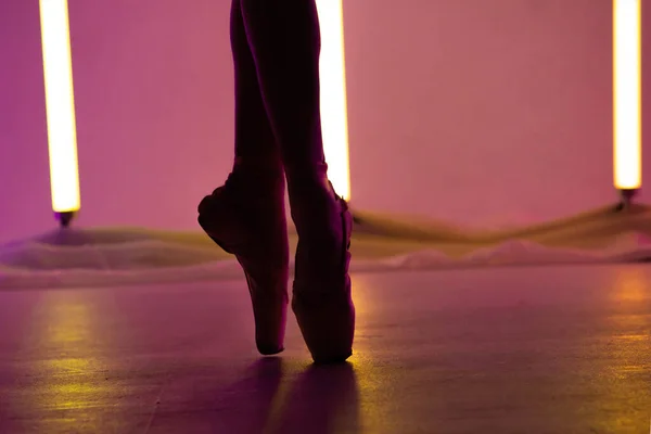 優雅なバレリーナは、明るいネオンの背景を背景に白い点の靴で古典的なバレエを踊っています。上のダンサーの細い足のシルエットを閉じる. — ストック写真