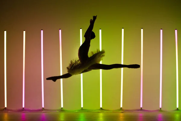 Bela jovem bailarina posando em um salto contra um fundo escuro estúdio com luzes de néon brilhantes. Silhueta de uma dançarina feminina delgada em um tutu branco e sapatos pontiagudos. — Fotografia de Stock