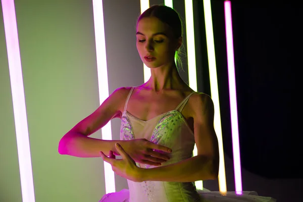 Retrato de uma bailarina em um tutu branco fazendo uma bela pose. Um dançarino de balé gracioso graciosamente posa contra o pano de fundo de luzes de néon brilhantes em um estúdio escuro. — Fotografia de Stock