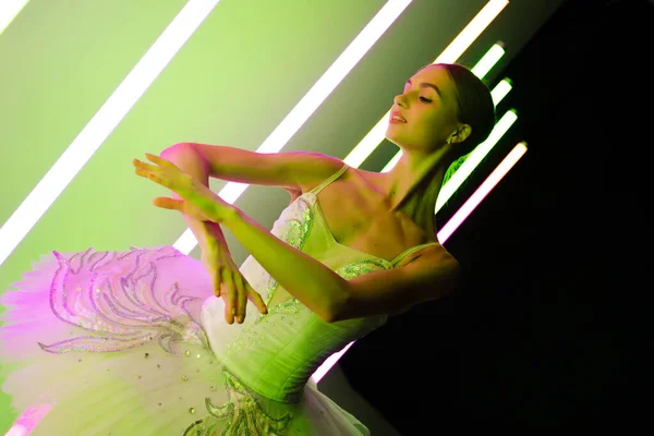 Retrato de una bailarina en un tutú blanco haciendo una pose hermosa. Una elegante bailarina de ballet posa graciosamente contra el telón de fondo de brillantes luces de neón en un estudio oscuro. —  Fotos de Stock