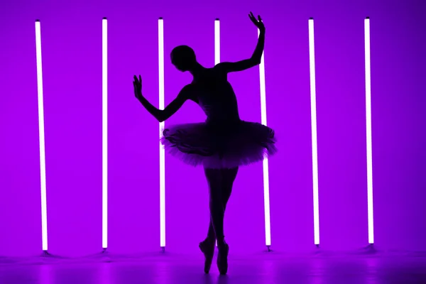 Young classical ballet dancer posing in a dark studio against a background of purple neon lights. Slender ballerina is silhouetted on tiptoes in white tutu. Ballet school poster. — Stock Photo, Image