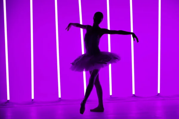 Young classical ballet dancer posing in a dark studio against a background of purple neon lights. Slender ballerina is silhouetted on tiptoes in white tutu. Ballet school poster. — Stock Photo, Image
