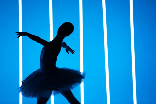Vacker ung ballerina dansar klassisk balett i en mörk studio mot en bakgrund av ljusa neonljus. Silhuett av en smal kvinnlig dansare i vit tutu och tåskor. — Stockfoto