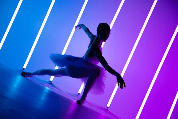 Νεαρή χαριτωμένη μπαλαρίνα σε pointe παπούτσια και ένα λευκό tutu δείχνει τις χορευτικές της ικανότητες. Μια όμορφη χορεύτρια κλασικού μπαλέτου με φόντο τα λαμπερά φώτα νέον σε ένα σκοτεινό στούντιο. Σιλουέτα. — Φωτογραφία Αρχείου