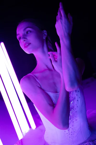 Porträtt av en ballerina i rollen som en vit svan mot bakgrund av starka neonljus i en mörk studio. Vacker ung modell graciöst poserar med händerna. — Stockfoto