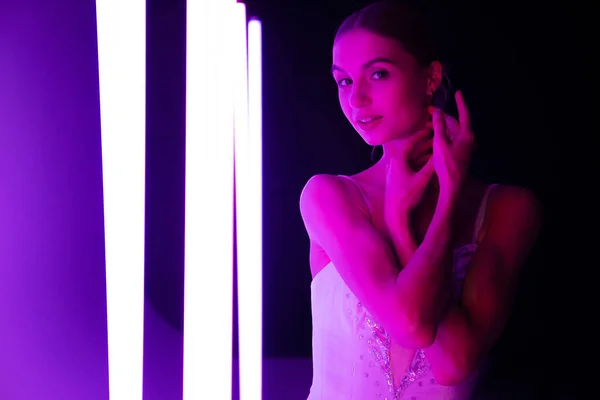 Retrato de uma bailarina no papel de um cisne branco contra um fundo de luzes de néon brilhantes em um estúdio escuro. Linda jovem modelo graciosamente posando com as mãos. — Fotografia de Stock