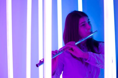 Genç ve güzel bir kadın flüt çalıyor. Parlak neon ışıkların arka planında karanlık bir stüdyoda poz veren müzisyen kadın. Kapatın. Bir müzik okulunun reklam afişi..