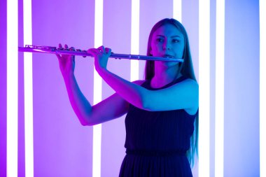 Siyah elbiseli genç ve güzel bir kadın flüt çalıyor. Parlak neon ışıkların arka planında karanlık bir stüdyoda poz veren müzisyen kadın. Kapatın. Bir müzik okulunun reklam afişi..