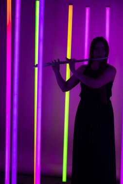 Siyah elbiseli genç ve güzel bir kadın flüt çalıyor. Parlak neon ışıkların arka planında karanlık bir stüdyoda poz veren müzisyen kadın. Siluet.