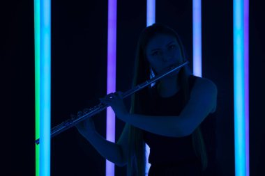 Kadın flüt çalıyor. Güzel flütçü bir rüzgar enstrümanı çalıyor. Parlak neon ışıklarının arka planında uzun saçlı güzel bir kadın manken. Siluet. Kapat..