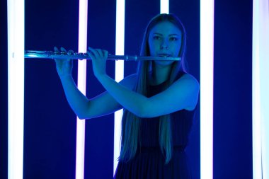 Kadın flüt çalıyor. Güzel flütçü bir rüzgar enstrümanı çalıyor. Parlak neon ışıklarının arka planında uzun saçlı güzel bir kadın manken. Kapat..