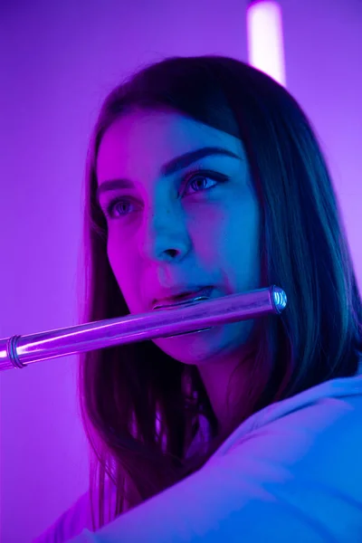 一个年轻美丽的女音乐家吹奏长笛的肖像。女性长笛手在明亮的霓虹灯背景下，在黑暗的工作室里演奏管乐器。靠近点. — 图库照片