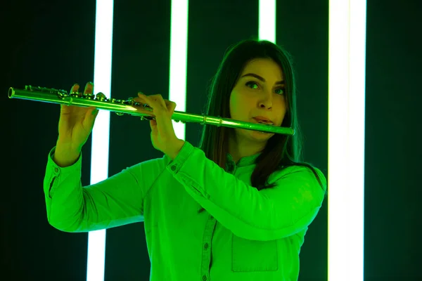 Woman in a white blouse plays the flute against a background of bright neon lights. Cute young female artist with a musical instrument in his hands. Poster for advertising a music school. Close up.