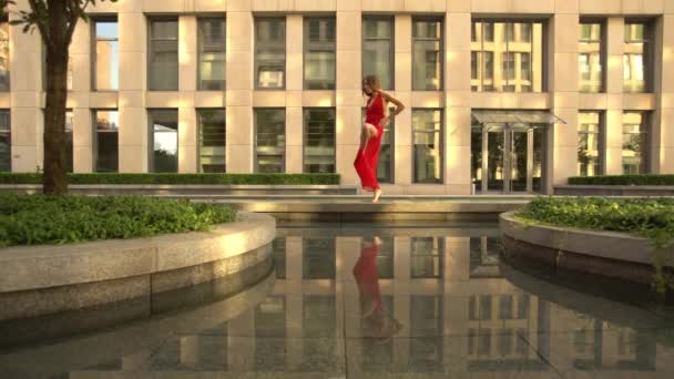 Beautiful young girl dancing on the street of a modern city and is reflected in the water. She is wearing a red dress. Slow motion — Stock Video