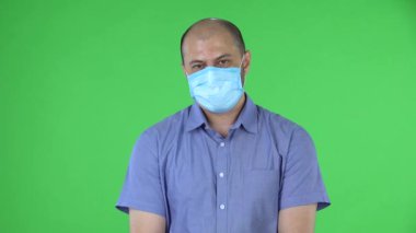 Medikal maskeli orta yaşlı bir adamın portresi baş sallamalarını altüst etti. Mavi gömlekli kel adam stüdyoda yeşil ekranda poz veriyor. Kapat..