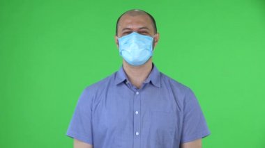 Medikal maske takmış orta yaşlı bir adamın portresi biriyle iletişim kuruyor ve onaylı bir şekilde başını sallıyor. Mavi gömlekli kel adam stüdyoda yeşil ekranda poz veriyor. Kapat..