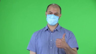 Medikal maskeli orta yaşlı bir adamın portresi baş parmak işareti gösteriyor. Mavi gömlekli kel adam stüdyoda yeşil ekranda poz veriyor. Kapat..