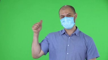 Medikal maskeli orta yaşlı bir adamın portresi yumruğunu gösteriyor, sonra bir, iki, üç, dört, beş parmak sayıyor. Mavi gömlekli kel adam stüdyoda yeşil ekranda poz veriyor. Kapat..