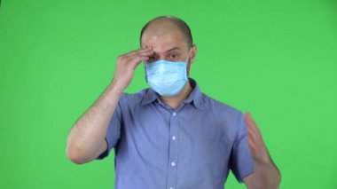 Medikal maskeli, endişeli ve üzgün orta yaşlı bir adamın portresi. Mavi gömlekli kel adam stüdyoda yeşil ekranda poz veriyor. Kapat..