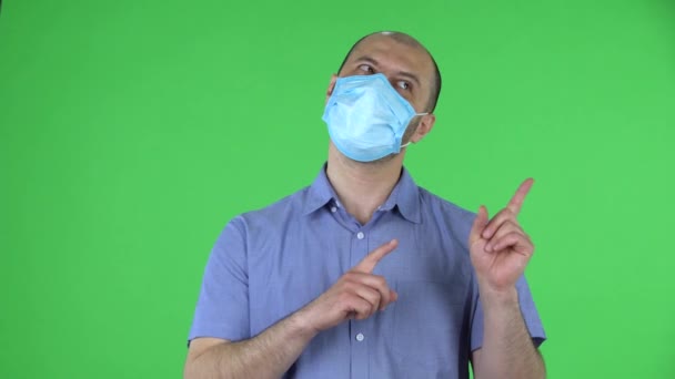 Portret van een man van middelbare leeftijd met een medisch masker wijst naar de zijkant. Kalende man in blauw shirt poseert op groen scherm in de studio. Sluiten.. — Stockvideo