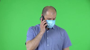 Medikal maskeli orta yaşlı bir adamın portresi akıllı telefonuyla konuşuyor. Mavi gömlekli kel adam stüdyoda yeşil ekranda poz veriyor. Kapat..